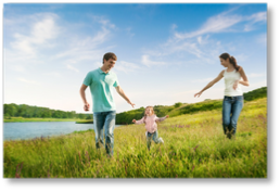 A couple run through wild grass next to a wide river, each stretching their hands out to their child running alongside between them and reaching for them both.