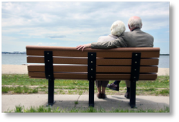 An older couple nestled together with his arm around her sit on a wooden bench overlooking a sandy beach.